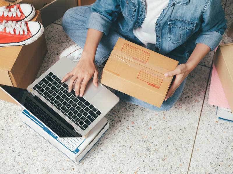 Woman holding a cardboard box and typing on a laptop